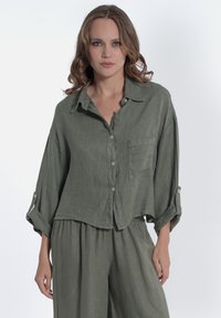 Camicia in lino verde oliva con bottoni, dalla vestibilità rilassata, tasca sul petto, maniche arrotolate e una texture sottile. Abbinata a pantaloni fluidi coordinati.