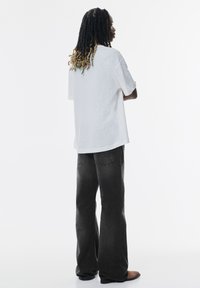 T-shirt bianco in cotone con una vestibilità rilassata, con maniche corte. Abbinato a jeans larghi grigi scuri. Scarpe piatte visibili.