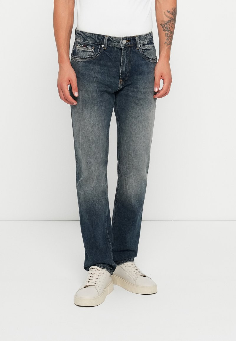 Homem a usar jeans retos pretos desbotados, tênis brancos e uma camisa branca, de pé contra um fundo branco liso.