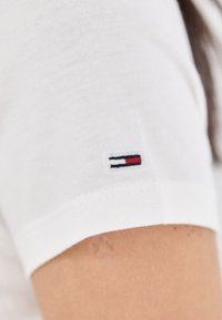 Baltas medvilninis marškinėlių rankovė su mažai siuvinėtu logotipu, kuriame yra raudonos, baltos ir mėlynos detalės, demonstruojanti klasikinį kasdienį dizainą.
