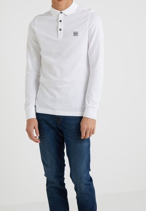 Weißes langärmliges Poloshirt mit Knopfleiste, mit einem kleinen grauen Logo-Patch und gerippten Bündchen. Kombiniert mit dunkelblauen Jeans.
