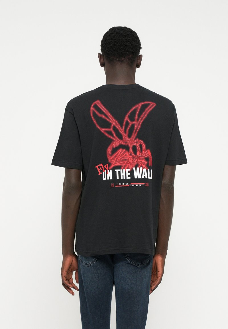 Schwarzes Baumwoll-T-Shirt mit einem roten Grafikmotiv einer Fliege mit großen Flügeln auf dem Rücken, darunter in Weiß der Schriftzug "Fly On The Wall".
