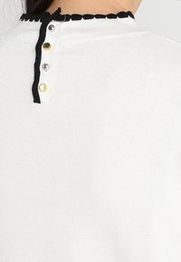 Haut en maille blanche avec bordure noire festonnée autour de l'encolure et quatre boutons-pression métalliques argentés et dorés dans le dos.