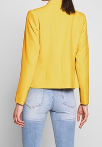 Blazer jaune sur mesure avec une texture lisse, une coupe ajustée et des manches longues. Porté avec un jean bleu clair déchiré.