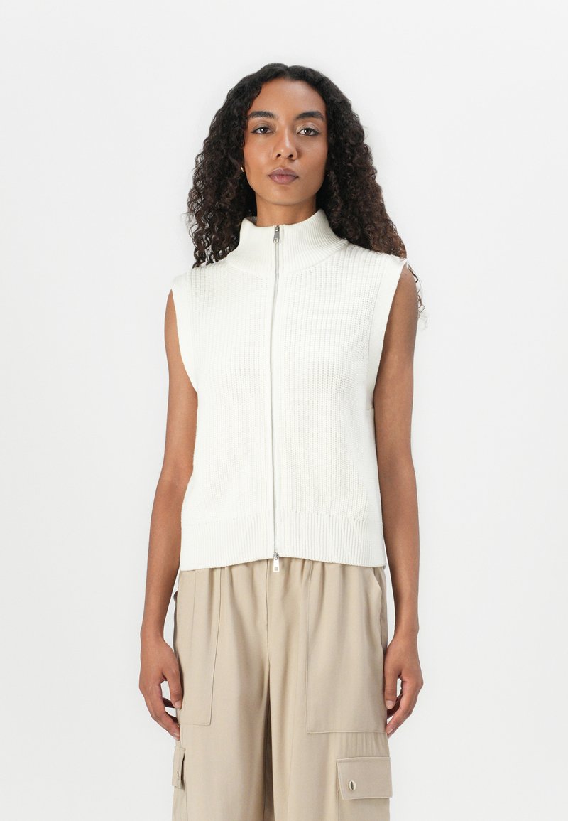 Débardeur en tricot sans manches crème avec un col haut et une fermeture éclair à l'avant, présentant un motif texturé et un ourlet côtelé. Idéal pour les tenues en couches.