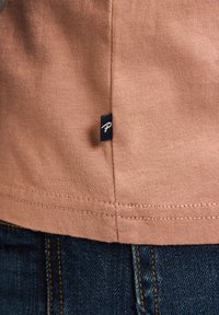 Světlé růžové bavlněné tričko s bočním štítkem v tmavě modré barvě. Disponuje pevným šitím a uvolněným střihem, v kombinaci s tmavě modrými džíny.