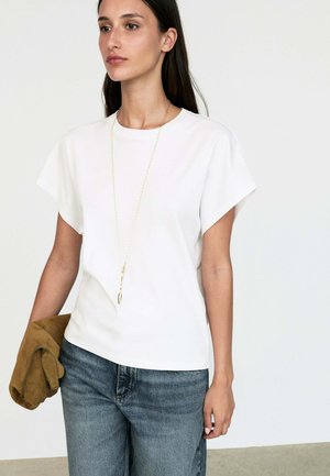 Femme portant un haut blanc uni à manches courtes, un jean bleu, un long collier en or, et tenant une veste marron pliée dans une main.