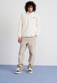 Pulover od flisa u boji off-white s četvrtim patentnim zatvaračem i logom na prsima, uparen s bež hlačama i smeškim tenisicama s bijelim detaljima.