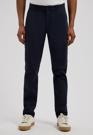 Man draagt zwarte slim-fit broek gecombineerd met witte sneakers met beige zolen tegen een egale lichte achtergrond.