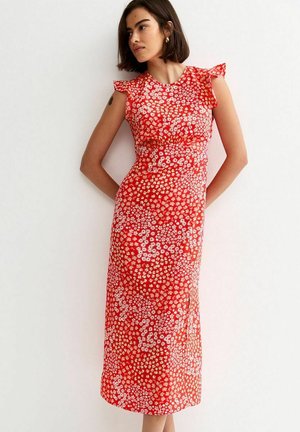 Vrouw met kort donker haar, gekleed in een mouwloze rode midi-jurk met een klein wit bloemenpatroon, staand tegen een witte muur met haar handen achter haar rug.