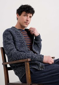 Navy en witte gestructureerde gebreide cardigan met lange mouwen en knopen, gedragen over een marineblauw en oranje gestreept shirt, zittend op een houten stoel.
