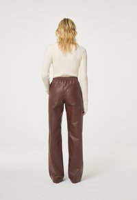 Bruine leren broek met hoge taille en elastische tailleband. Getextureerde afwerking en wijd uitlopende pijpen. Model draagt een crèmekleurige geribbelde top met lange mouwen.