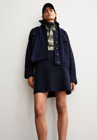 Marineblaue Wollmischjacke mit großen Knöpfen, kombiniert mit einem karierten Hemd. Verfügt über einen gerippten Kragen und einen passenden Rock mit einem strukturierten Muster.