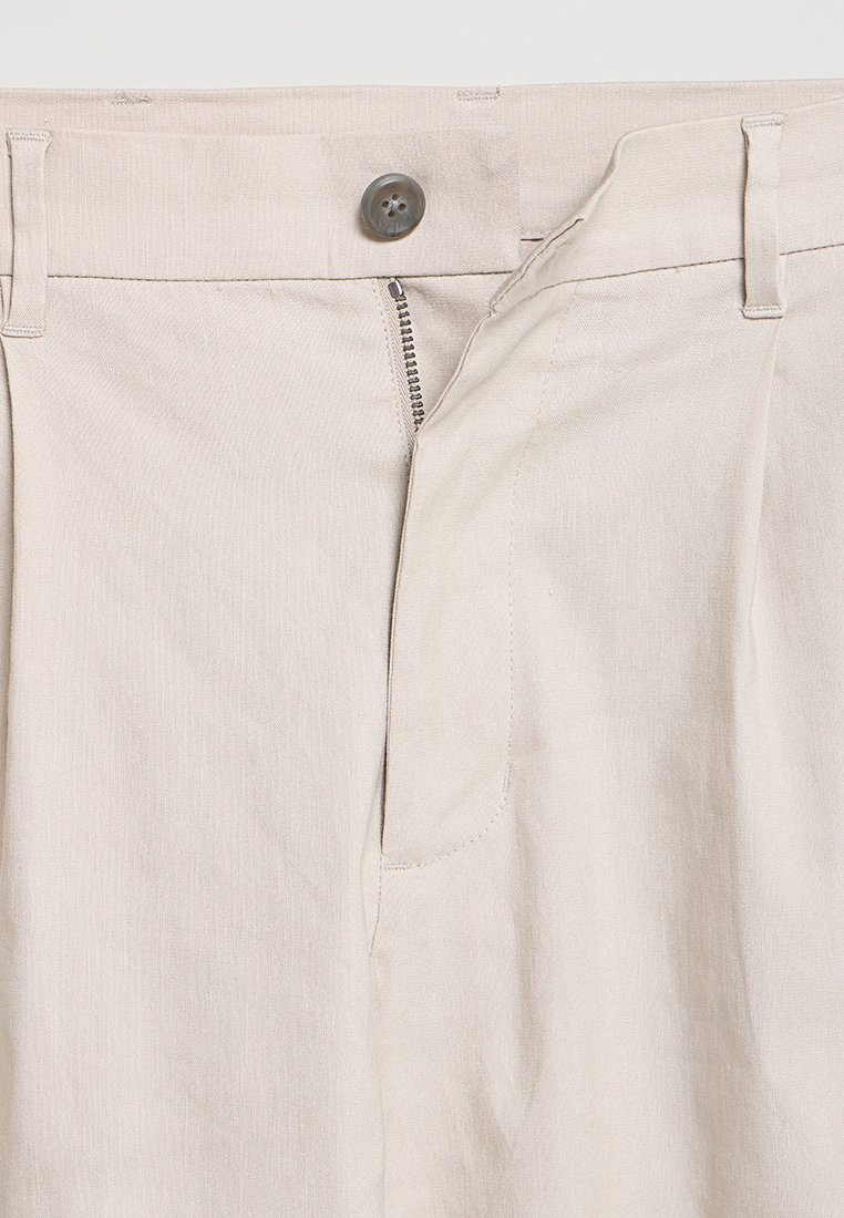 Beige Hosen mit teilweise geöffnetem Reißverschluss und Knopfverschluss, sichtbaren Gürtelschlaufen und einer sauberen, glatten Stoffoberfläche.