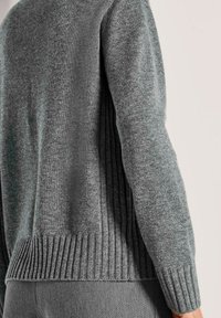 Nahaufnahme einer Person, die einen grauen Strickpullover mit gerippten Details am Saum, den Bündchen und den Seitenpaneelen trägt.