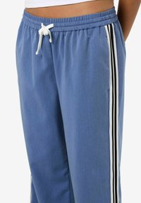 Un pantalon de survêtement bleu clair présentant une taille élastique avec cordon de serrage, des poches latérales et des rayures verticales noires et blanches le long des coutures extérieures.