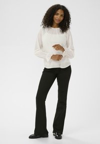 Blouse blanche légère avec de longues manches bouffantes et des poignets froncés, associée à un pantalon noir évasé et des chaussures noires brillantes.