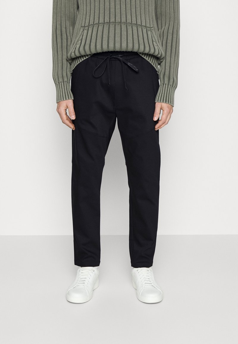 Personne portant un pantalon noir à cordon, un pull côtelé vert et des baskets blanches, debout devant un fond clair uni.