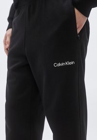 Calças de treino pretas com textura suave, apresentando um bolso lateral com fecho zip e o logótipo branco "Calvin Klein" na coxa. Design simples e corte reto.