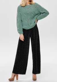 Hellgrüner gestrickter Pullover mit Wellenmuster, kombiniert mit schwarzen weit geschnittenen Hosen mit weißen Punkten und beigen klobigen Absätzen.