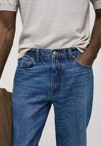 Jeans de mezclilla azul con corte recto, diseño de cinco bolsillos, costuras en contraste y cierre con botón en la cintura.