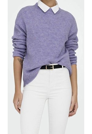 Personne portant un pull côtelé lavande par-dessus une chemise blanche à col, rentrée dans un pantalon blanc avec une ceinture noire.
