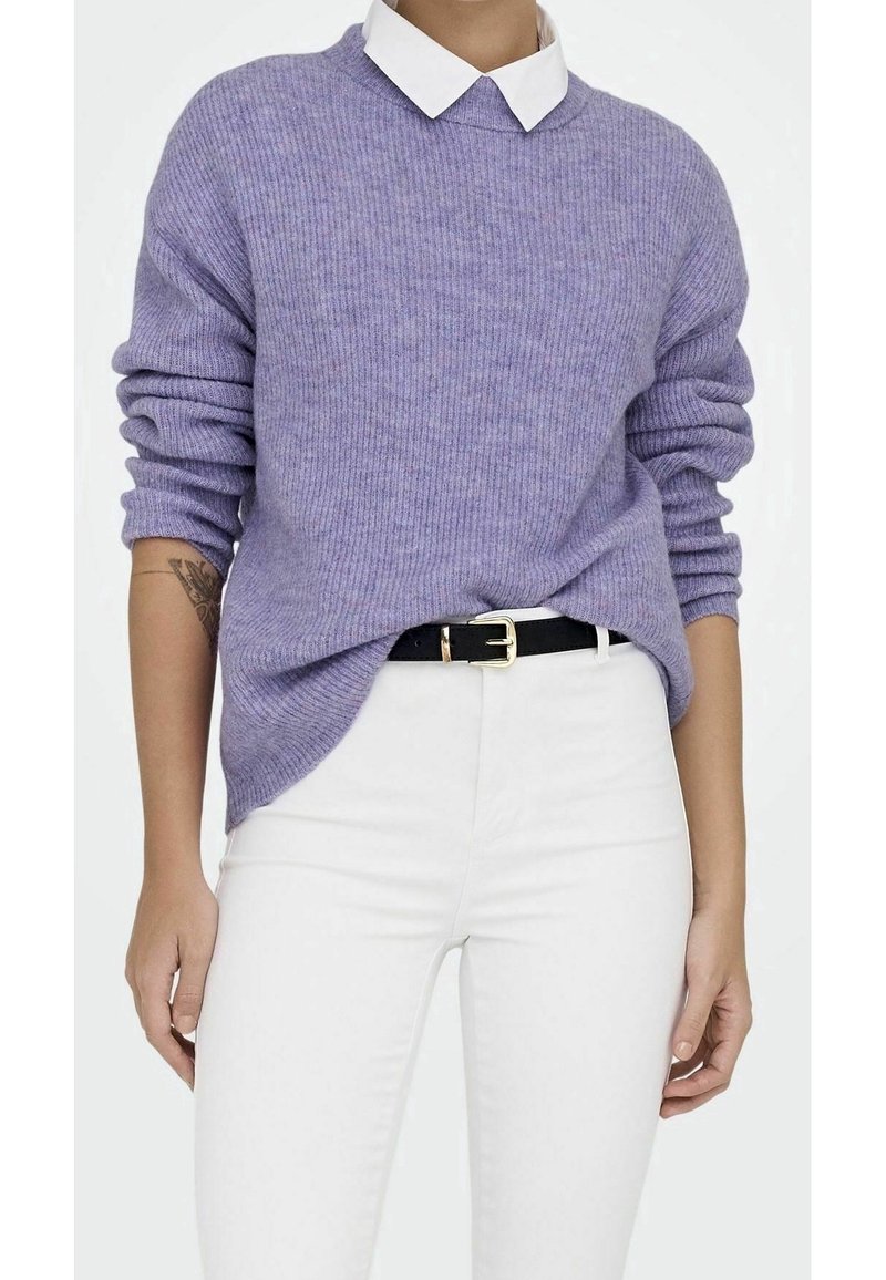 Personne portant un pull côtelé lavande par-dessus une chemise blanche à col, rentrée dans un pantalon blanc avec une ceinture noire.