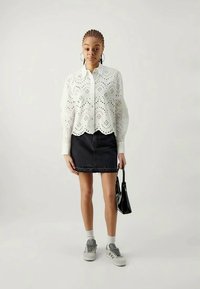 White lace shirt with cut-out patterns, black denim skirt, gray sneakers, and a small black handbag. Model stands against a light background.
