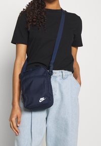 Tmavě modrá taška na rameno Nike s obdélníkovým tvarem, syntetický materiál, bílé logo, nastavitelný popruh a kapsy na zip.