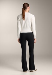 Bela pletena jopica z dolgimi rokavi in ribastim robom, v kombinaciji z črnimi širokimi hlačami. Ozadje je nevtralno in nekonfliktno.