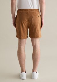 Pantalones cortos de tela marrón y suave con cinturilla elástica, que cuentan con un único bolsillo trasero y un corte holgado, combinados con zapatillas blancas.