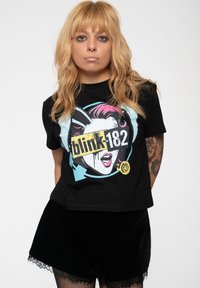 Black cotton t-shirt featuring a graphic print of a face with pink hair and the text "blink-182" in yellow. Worn with black velvet shorts.