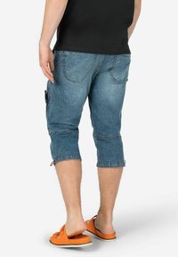 Jeans-Cargo-Shorts in Hellblau, über dem Knie gekürzt, mit seitlichen Taschen und dekorativen Nähten. Tragen mit orangefarbenen Slipper-Sandalen.