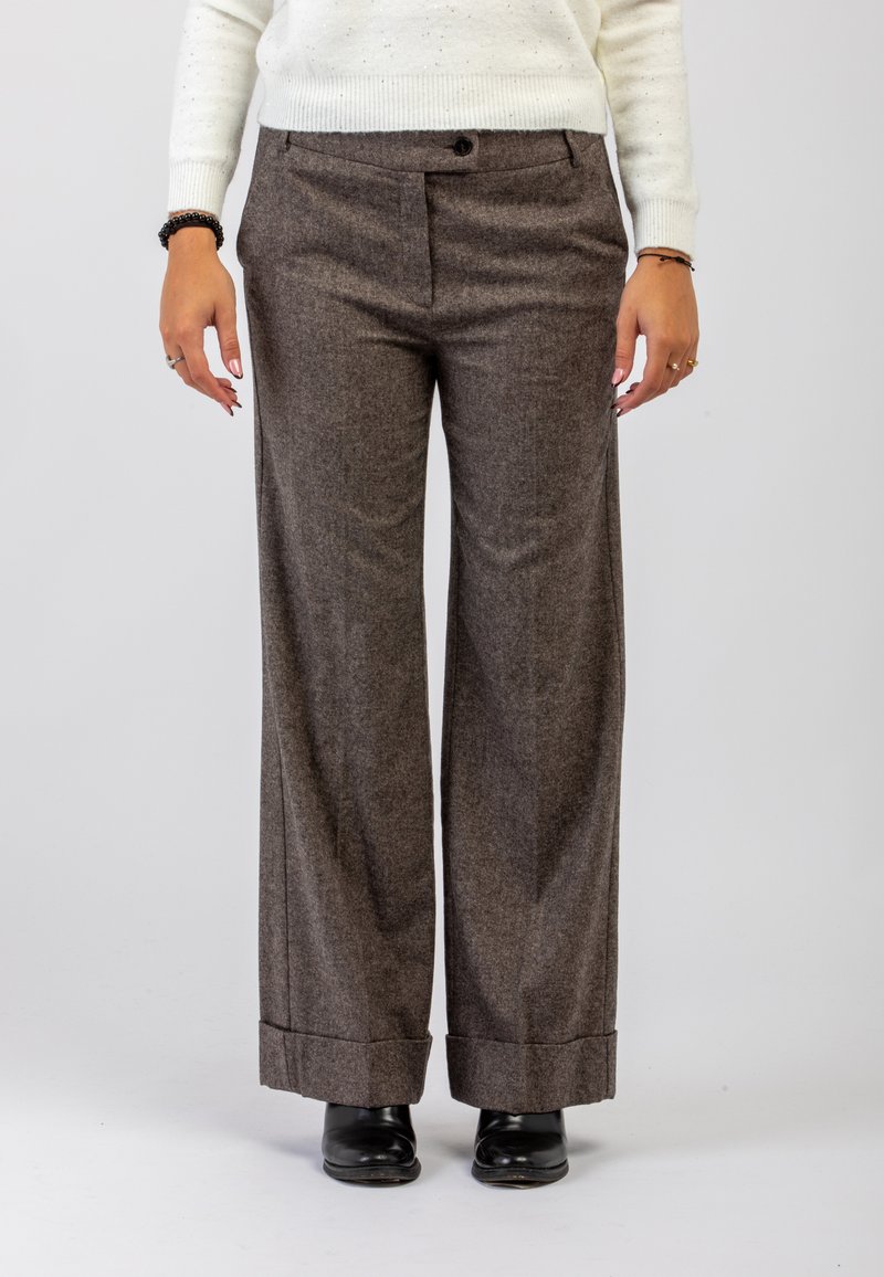 Persona che indossa pantaloni larghi marroni, stivali neri e un maglione di maglia bianco, in piedi contro uno sfondo chiaro e semplice.