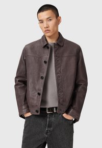Homme portant une veste en cuir marron foncé par-dessus un pull gris clair, avec les mains dans les poches d'un jean noir, se tenant devant un fond gris uni.