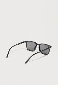 Μαύρα ορθογώνια γυαλιά ηλίου με σκουρόχρωμους φακούς και λεπτούς βραχίονες, ανοιχτά τοποθετημένα σε φωτεινή επιφάνεια.