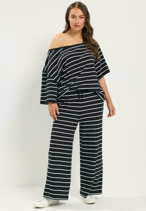 Femme portant un pantalon large rayé horizontal noir et blanc et un top épaules dénudées, associé à des baskets blanches, debout devant un fond uni.