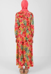 Maxi mekko pitkillä hihoilla, kukkakuosi punaisena ja vihreänä keltaisella taustalla, vyötetty vyötäröltä, ja siihen sopiva huivi.