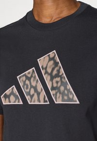 Črna bombažna majica s kratkimi rokavi, ki prikazuje velik, stiliziran adidasov logotip z vzorcem leoparda, obrobnjenim z svetlo roza barvo. Okrugli ovratnik.