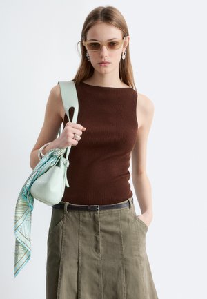 Femme portant un haut marron sans manches et une jupe olive, tenant un sac à main vert clair avec un foulard à motifs, portant des lunettes de soleil et des boucles d'oreilles argentées.