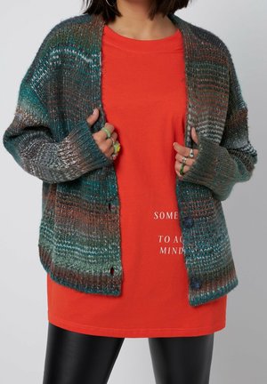 Personne portant un cardigan en tricot multicolore sur une chemise orange avec texte blanc partiel, tenant les bords du cardigan, et un pantalon en cuir noir.