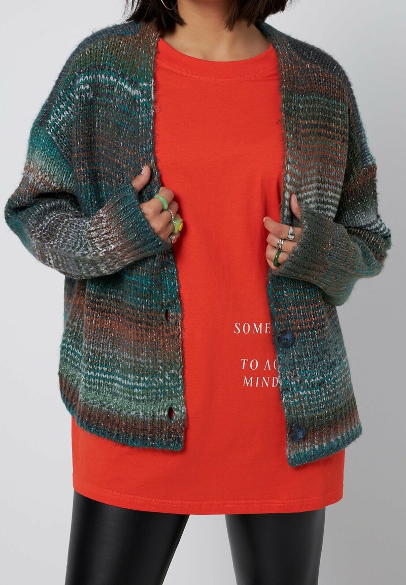 Personne portant un cardigan en tricot multicolore sur une chemise orange avec texte blanc partiel, tenant les bords du cardigan, et un pantalon en cuir noir.
