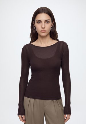 Femme aux longs cheveux bruns, portant un haut côtelé marron foncé ajusté à manches longues et un pantalon plissé marron clair, debout sur un fond uni.