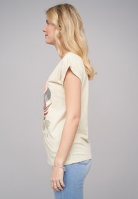 T-shirt beige clair avec un design graphique floral, manches courtes retroussées et coupe décontractée, associé à un jean bleu.