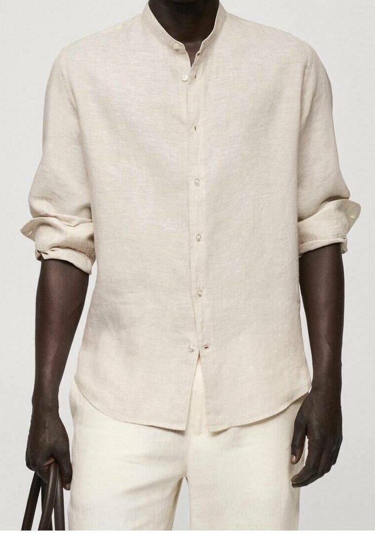 Homme portant une chemise en lin beige à col mandarin et un pantalon assorti, tenant un sac brun foncé, manches retroussées.