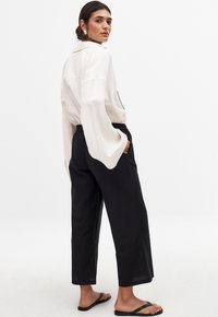 Crème oversized blouse met brede mouwen, gecombineerd met zwarte losse broeken. Het model draagt zwarte slippers en heeft een minimalistische oorbel.