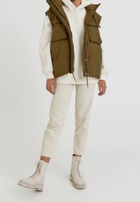 Khakifarbenes Puffer-Weste mit großen Taschen, übereinander getragen mit einem cremefarbenen Kapuzenpullover, kombiniert mit off-white gerippten Hosen und hellbeigen Schnürstiefeln.