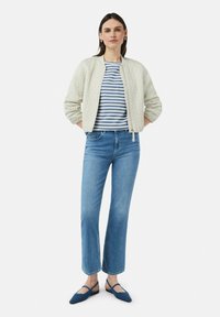 Veste bomber matelassée de couleur claire, haut rayé bleu marine et blanc, jean bleu et ballerines bleues avec des lanières. Textures lisses et coupe décontractée.
