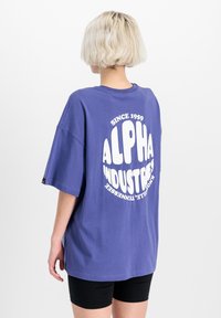 Fioletowy, oversized t-shirt z okrągłym białym logo na plecach, które wytłoczonymi literami głosi "ALPHA INDUSTRIES SINCE 1959".