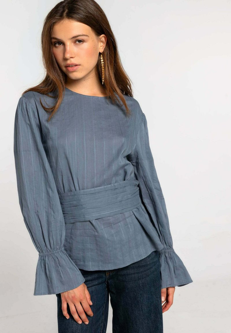 Blusa de rayas azul claro, con escote redondo, cintura atada y mangas acampanadas. Combinada con jeans de mezclilla oscuros.